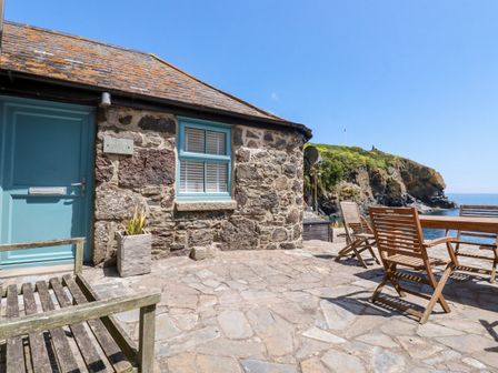 An outdoor patio with wooden chairs and a table next to a stone cottage by the sea at Beach Cottage in Cadgwith