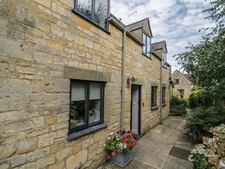 The exterior of a stone cottage with windows and a door along a paved pathway with flowers and bushes at The Cottage in Chipping Campden