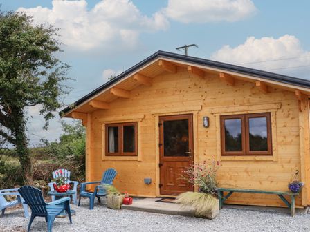 A wooden cabin with a brown door and windows with blue chairs and a green bench outside surrounded by plants and gravel at Wheatfield Cabin in Ramstown near Fethard-On-Sea County Wexford