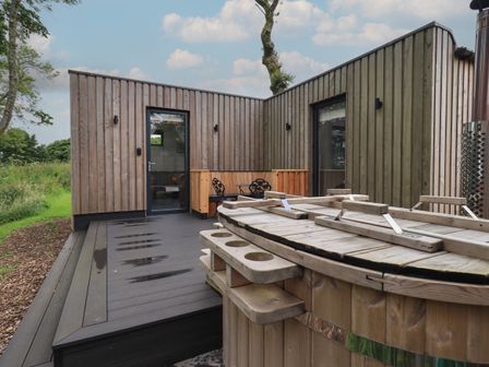 An outdoor wooden deck with a hot tub a seating area and two glass doors at Bluebell Cabin in Ullswater