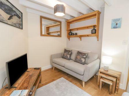 A small living room with a gray sofa two pillows a wooden shelf with decor a mirror a wooden TV stand a TV a small wooden nesting table with a lamp and a framed print on the wall at 5 Tai Isa in Barmouth