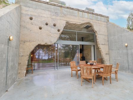 An outdoor patio area with a wooden table and six wooden chairs in front of a modern concrete structure with large sliding glass doors at Standby Generator Bunker Ringstead near Weymouth