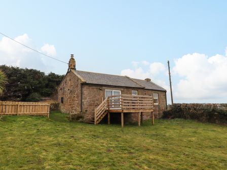 A stone cottage with a small wooden deck and grassy yard at Henhill Lodge in Chathill near Seahouses
