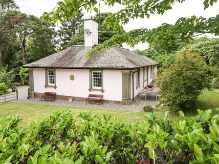 A single-story house with two benches and a table set outside surrounded by green trees and bushes at Upper Lodge in Bowness-On-Windermere