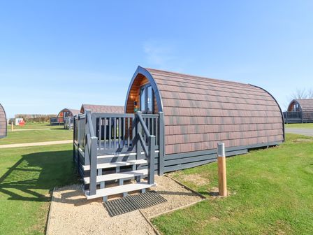 A small wooden pod with stairs and a railing on a grassy area at Owl's Nook in Kilkhampton