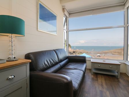 A sitting area with a black leather sofa a small table with drawers a lamp and a large window showing a beach at At the Bay Apartment in Hele Bay Ilfracombe