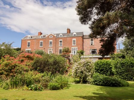 A large brick house with multiple windows behind a garden with bushes flowers and trees at Rock House Apartment in Llanllwchaiarn near Newtown