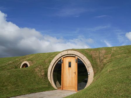 A round wooden door built into a grassy hillside under a blue sky at Badgers View in Rhayader