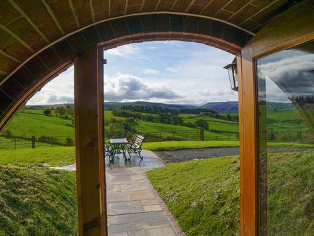 A curved wooden door opening to a stone patio with a table and chairs overlooking green hills at Foxes View in Rhayader
