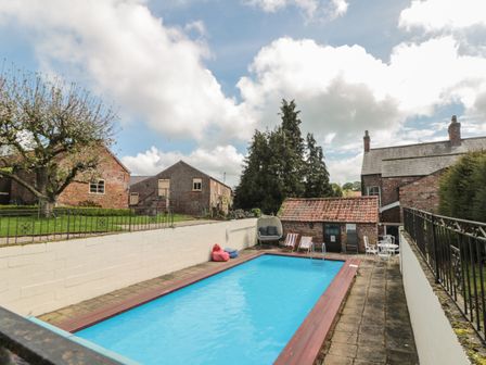 An outdoor swimming pool with deck chairs bean bags and a hanging chair surrounded by brick buildings at Bailey house in Pocklington