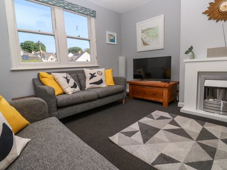 A living room with grey sofas and patterned cushions a wooden TV stand with a television a geometric patterned rug and a fireplace at 8 Rosewall Cottages in St Ives