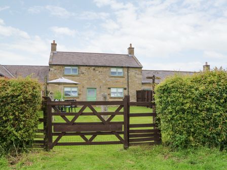 A stone house with green framed windows and door behind a wooden gate and garden at The Granary in Longwitton near Morpeth