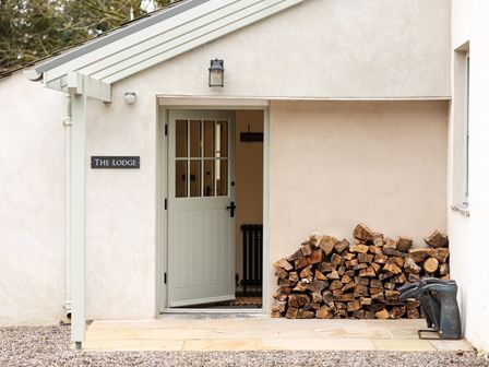 Front door entrance to a building with a pile of firewood and rubber boots outside at The Lodge in Newby Bridge