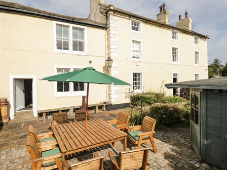 An outdoor patio with a wooden table and chairs under a green umbrella next to a light yellow building and a small green shed at Abbey Farm House in St Bees