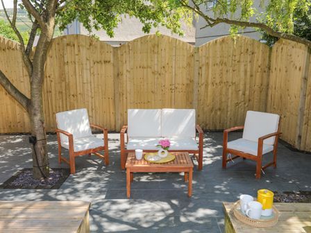An outdoor seating area with wooden chairs and a table under a tree in a fenced yard at Maes Y Ffynnon in Nefyn