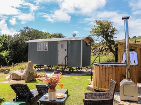A shepherds hut with wheels and a hot tub with chimney on grass with chairs and a table with flowers at Bosulla Shepherds Hut in Penzance