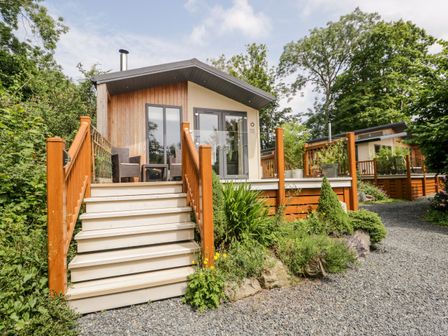 A small wooden cabin with stairs leading to a porch and outdoor chairs at Pippin Lodge in Allithwaite near Cartmel