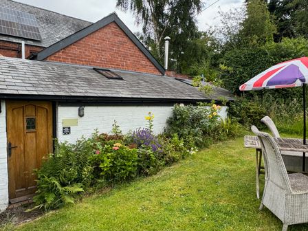 A garden with flowers and plants next to a white brick building with a wooden door and table with chairs and an umbrella at The Old Bakehouse in Pembridge