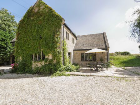 A stone house with ivy on the wall a patio with table and umbrella and surrounding gravel and grass at Paddock Barn in Castle Combe