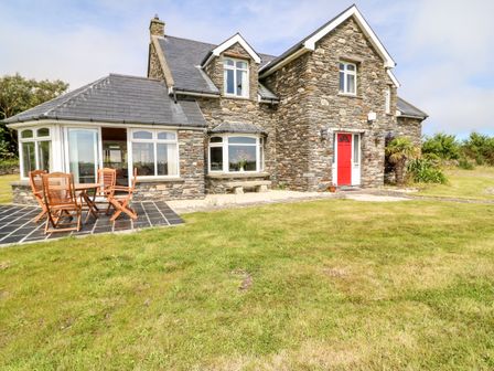 A stone house with a red door and patio furniture on a tiled area in front of a grass lawn at Coorlacka Goleen County Cork