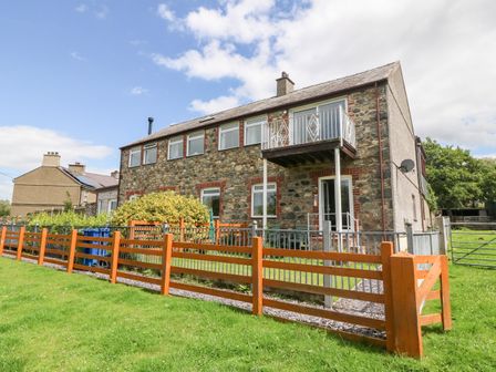 Exterior view of a stone house with multiple windows a small balcony and a wooden fence surrounding the grassy yard at Coed y Glyn Bach Equestrian in Penygroes