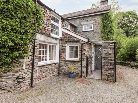 An exterior view of a stone cottage with white framed windows ivy on the wall and a gravel driveway at Holly Cottage in Grasmere