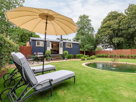 A garden with two lounge chairs under an umbrella a round pond and a small dark blue house with open doors at Utopia in Metfield near Harleston