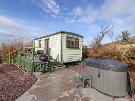 An outdoor area with a small green cabin with a deck and outdoor furniture a charcoal grill and a covered hot tub at Nyth Y Bioden Magpies Nest Llanaelhaearn near Trefor