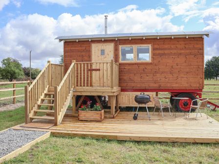 A wooden shepherds hut on wheels with wooden stairs and a small deck with a barbecue grill two chairs and a table at Shepherds Hut in Garthorpe near Scunthorpe