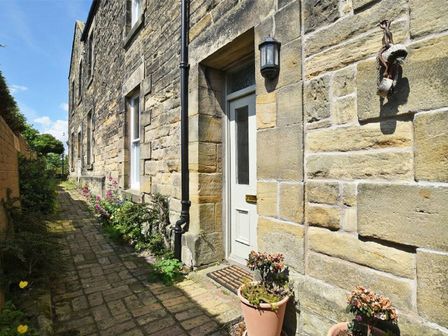 A stone exterior wall with a white door and window along a brick pathway with potted plants and flowers at Riverbank Cottage in Alnmouth
