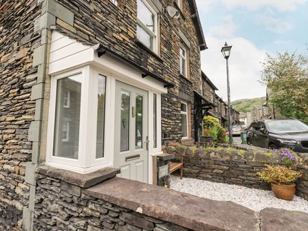 Stone house exterior with white door and windows a small garden with a bench and potted plant on a stone wall at Woolly End Cottage in Ambleside