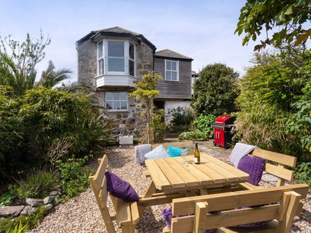An outdoor garden area with a wooden picnic table and benches in front of a stone house with greenery and a red barbecue at Ayr Farmhouse in St Ives
