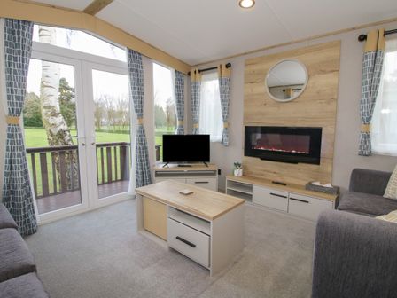 A living room with sofas a coffee table a TV an electric fireplace and large windows with curtains at Heron in Shobdon