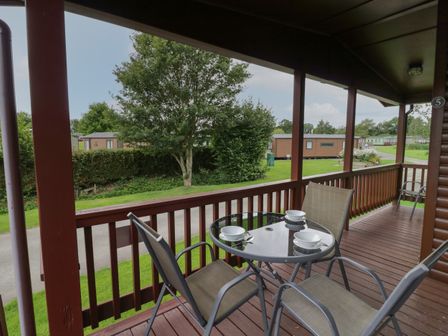 A covered wooden porch with a glass table and four chairs overlooking a grassy area with trees and mobile homes at Daffodil in Eardisland