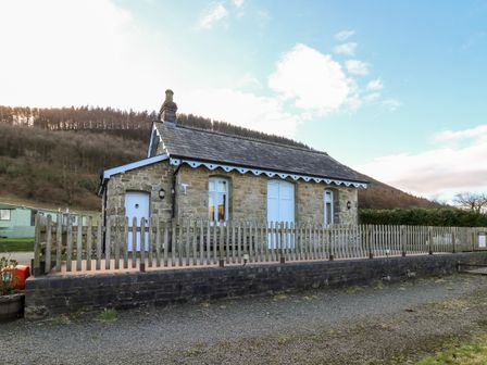 A stone building with a wooden fence and a gravel pathway in front of hills at Railway Station Cottage in New Radnor near Kington