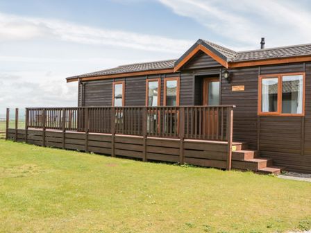 A wooden lodge with multiple windows and a railing on a deck in a grassy area at Three Views Lodge Whitsand Bay Fort Holiday Village