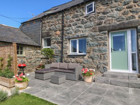 An outdoor patio with gray tile flooring a gray sectional sofa a small table red grill potted flowers and a stone building with a green door at Yr Ysgubor in Dyffryn Ardudwy