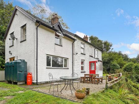 A white two-story house with a red door a small patio with metal and wooden furniture and a green fuel tank at Delfryn Goginan in Goginan near Aberystwyth