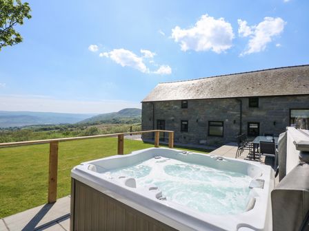 An outdoor patio with a hot tub and dining table next to a stone building with a view of hills and sky at Ysgubor Maen Y Bardd in Rowen near Conwy