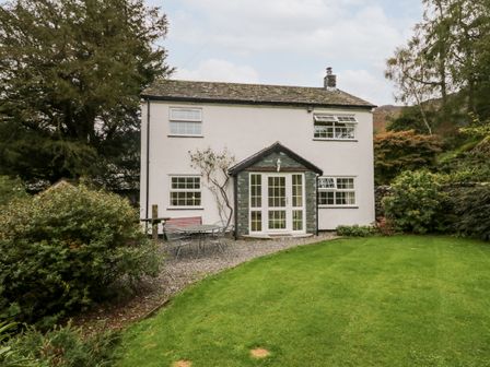 A white two-story house with a small porch and garden furniture on a gravel area in front of a green lawn at Thwaite How in Borrowdale