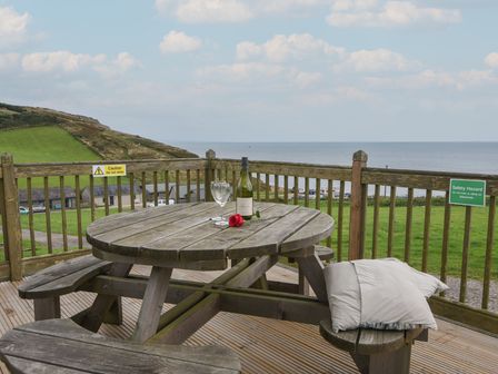 A wooden picnic table with two pillows a wine bottle two glasses and a red rose on a wooden deck overlooking a grassy hill and the sea at Sealark in Eype