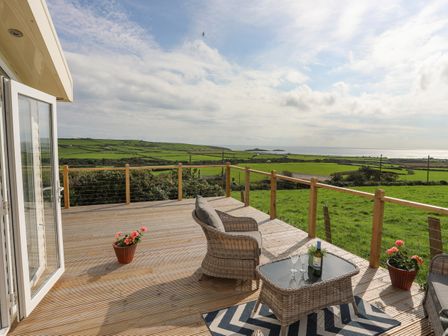 An outdoor deck with wicker chairs table and flowers overlooking green fields and the sea at Cae'r Wylan in Aberdaron