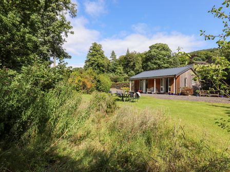 A wooden cabin with outdoor furniture on a grassy lawn surrounded by trees at Watersong in Coniston