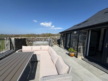 An outdoor patio with a sectional sofa and table next to a stone building and a stone wall with a hot tub behind Gaerau Bach in Llanaber