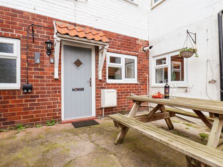 An outdoor patio with a wooden picnic table and benches a bottle of wine and glasses on the table a gray door with a small roof above it and several white framed windows at 4 Boulby Bank Top Whitby