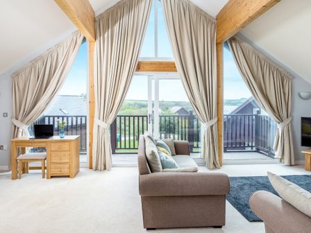 A living room with large windows and beige curtains a brown sofa a wooden desk with a chair and laptop and a balcony at Sunbow at Retallack Resort in Winnards Perch St Columb Major