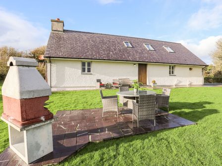 An outdoor patio with a table and chairs next to a barbecue grill in front of a house with skylights and a wooden door at Rhyd Y Bont Bach in Rhoscolyn