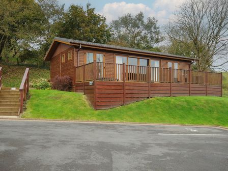 A wooden lodge with a fenced porch on a grassy hill near a parking area at Hideaway Lodge Bossiney near Tintagel