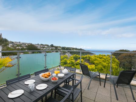 An outdoor dining table set with plates, cups, croissants, strawberries, and juice on a terrace overlooking houses and a coastal view at 7 Sandy Lane in Carbis Bay