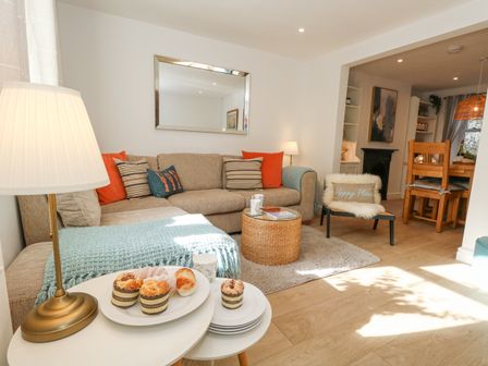 A living room with a beige sectional sofa and colorful cushions a wicker coffee table a small round white table with baked goods a lamp a chair with a pillow a dining area with wooden chairs and a table at Winkle Cottage in Weymouth